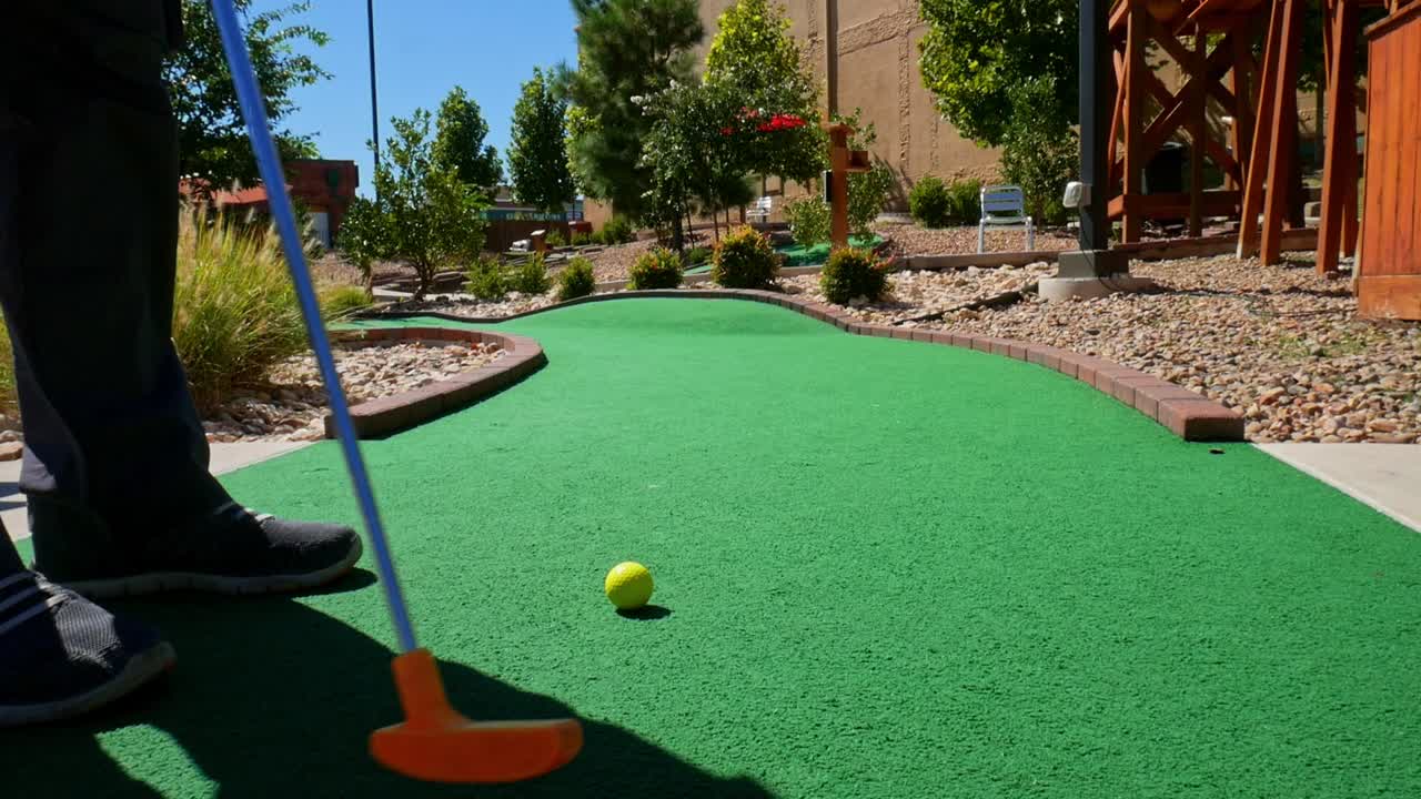 Playing mini golf with a yellow ball and orange putter on green astroturn on a sunny day - shot from behind golfer