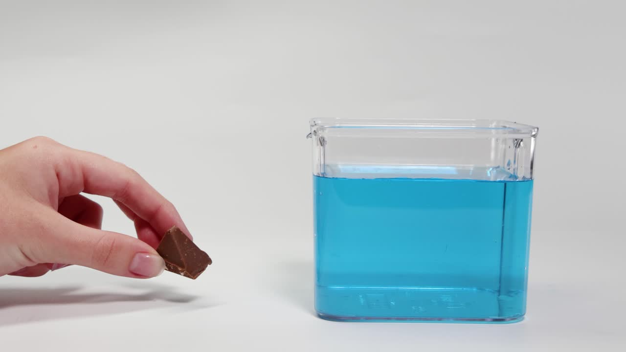 A hand drops a chocolate piece into a clear container of blue liquid, observing its sinking behavior