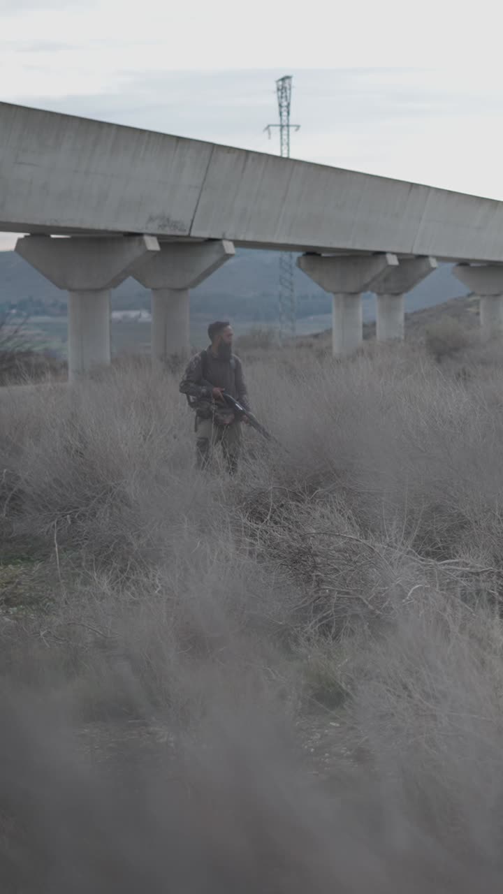 Man with rifle in field under bridge