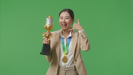 asian business woman in a suit with a gold medal and trophy showing call me gesture and smiling to camera as the first winner on green screen background in the studio