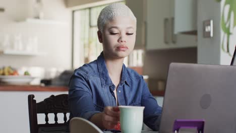 mujer biracial ocupada trabajando en casa con una computadora portátil y bebiendo café en cámara lenta