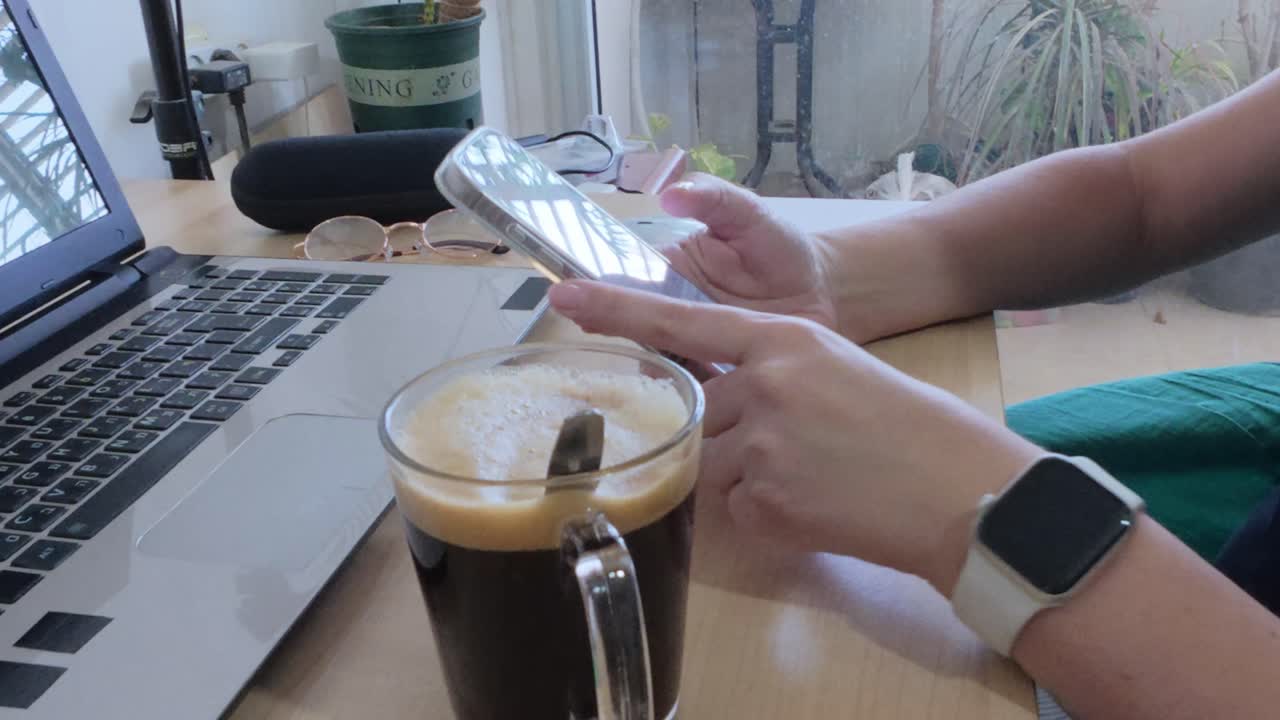 Person enjoying coffee break while using phone at desk with laptop.