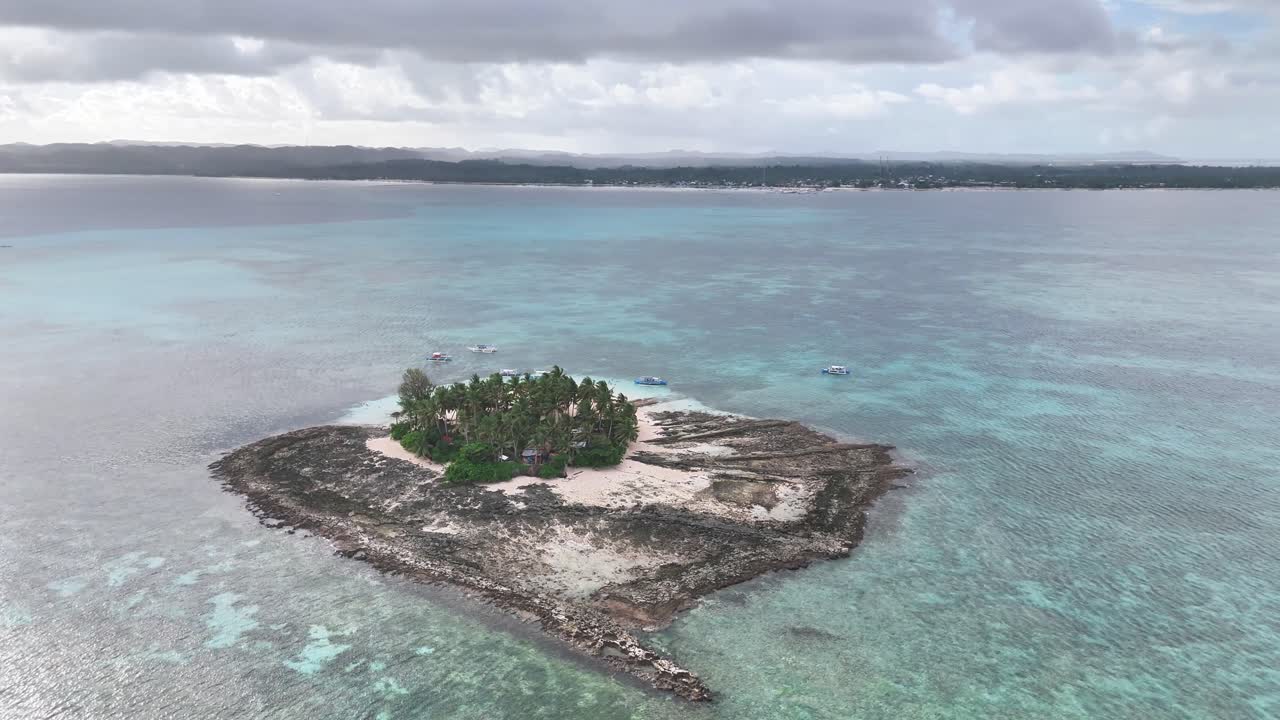 Aerial view of a small tropical island near Guyam Island, Siargao, surrounded by turquoise waters, coral reef, and palm trees under a partly cloudy sky with boats anchored nearby