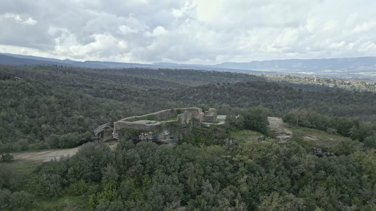 Lush green forest surrounds the ancient castle of taradell, built atop a rocky hill, showcasing its historical architecture against the backdrop of a cloudy sky