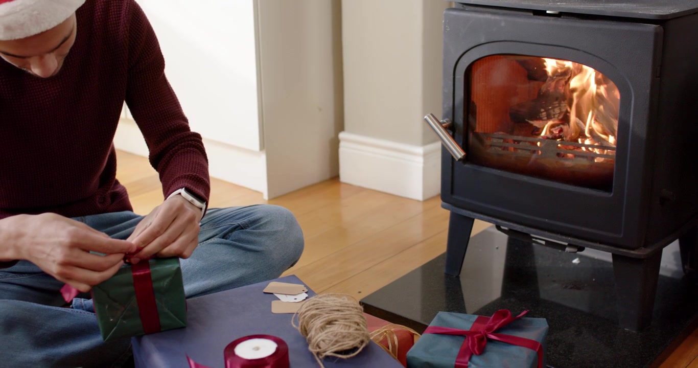 Uomo seduto accanto alla stufa a legna che lega un nastro rosso su una scatola verde, indossando un cappello di Babbo Natale, preparando i regali.