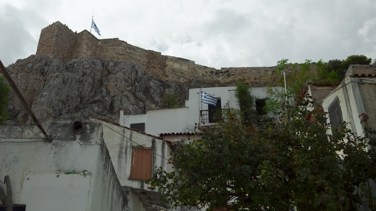 Vista de baixo para cima da bandeira grega no topo da Acrópole e casas típicas.