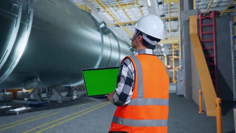 vista trasera de un ingeniero masculino asiático con casco de seguridad de pie en una fábrica de fabricación de tubos. mirando una computadora portátil de pantalla verde y mirando a su alrededor