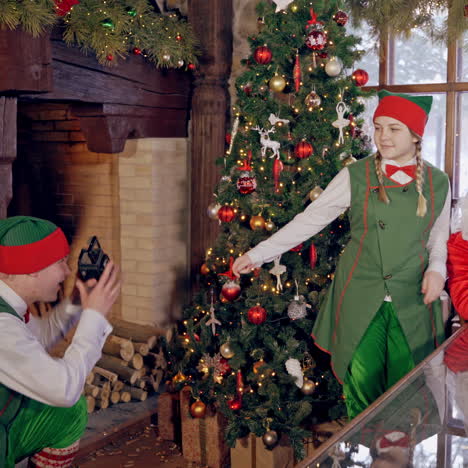 happy elf and santa near the christmas tree. elf is taking photo of a happy fairy characters in decorated room.