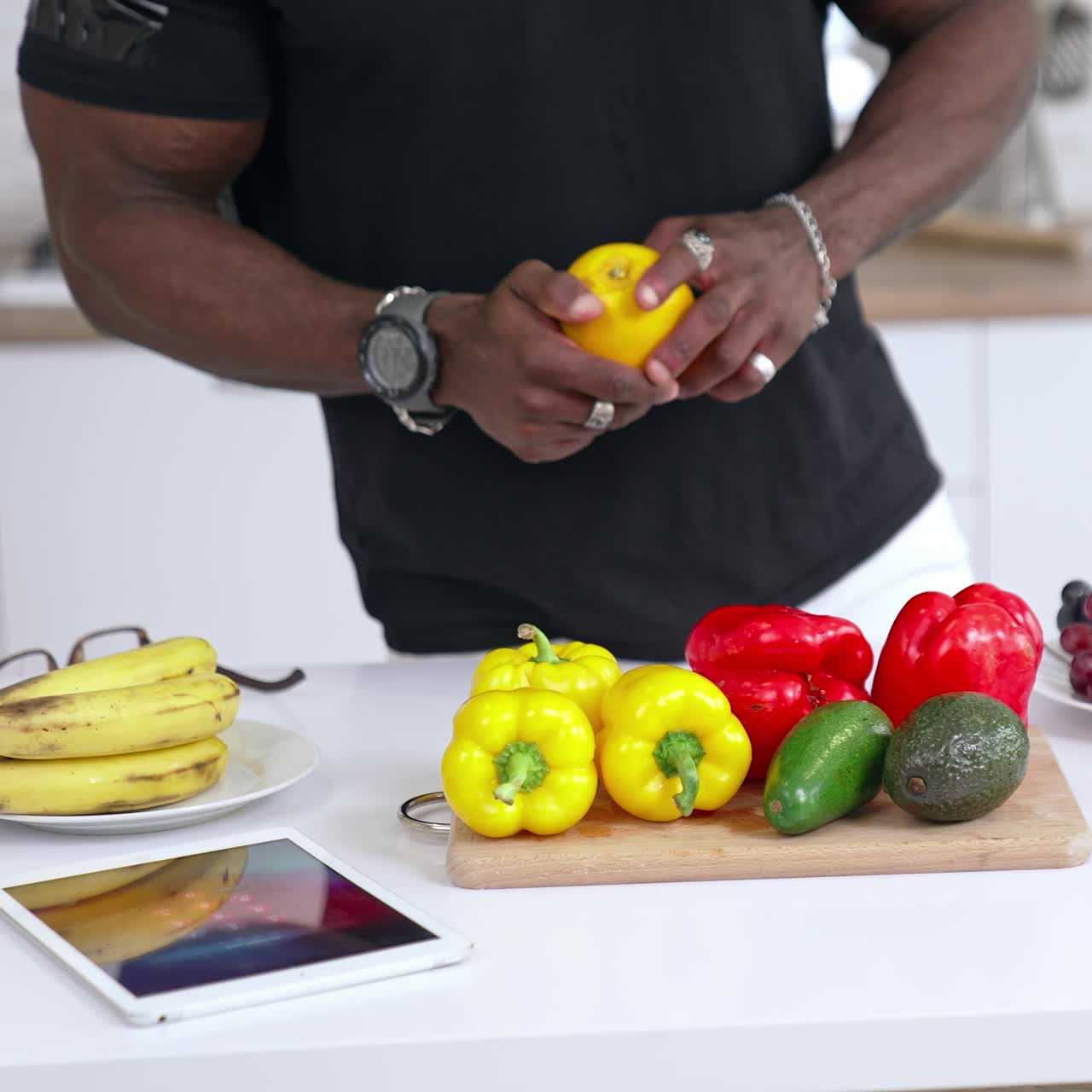 Multiracial man standing at the kitchen full of fruits and vegetables in the modern interior and looking at the tablet. Food and health care concept