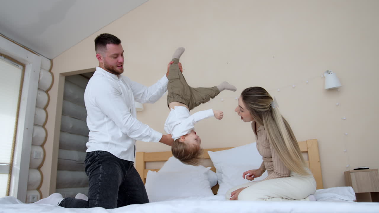 Father turns and holds his baby upside down. Parents spend fun active time in the bedroom.