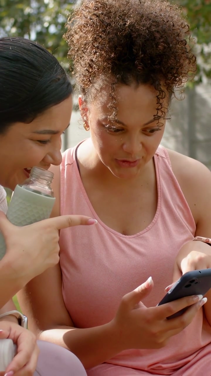 Vertical video: Pink-top woman holding phone, friend offering cap starting social media alerts
