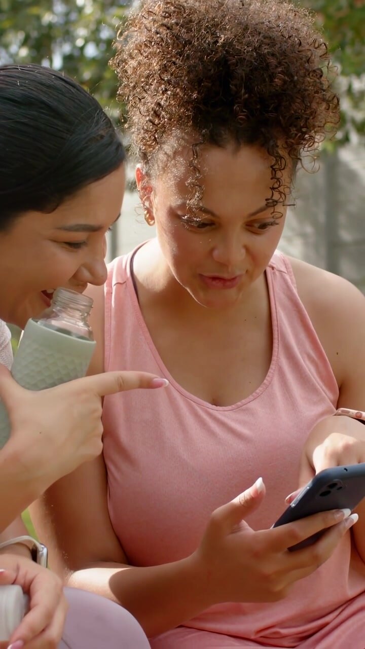 Vertical video: Pink-top woman holding phone, friend offering cap starting social media alerts