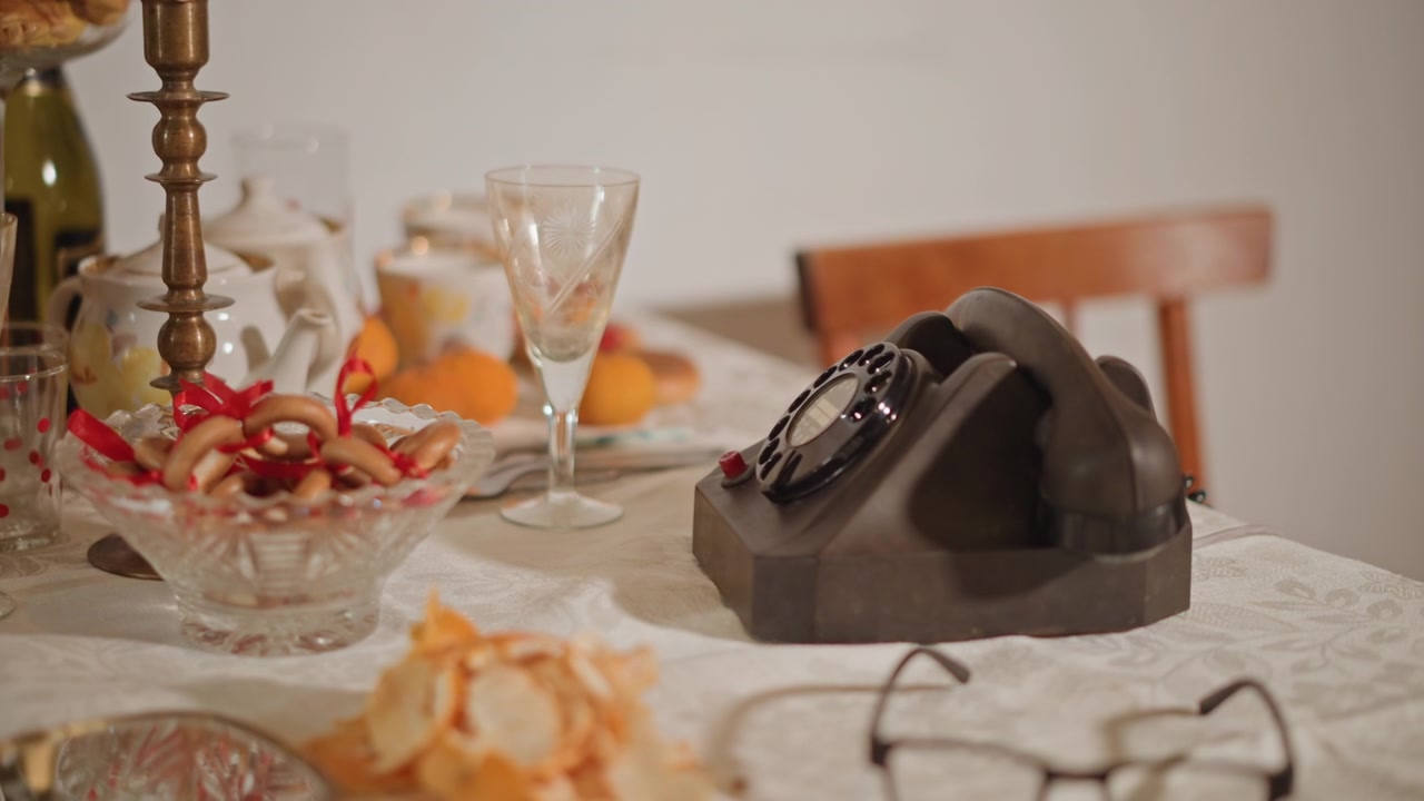Vintage rotary phone on table, soft warm light, lace tablecloth, glassware and candy bowl, brass candlestick, empty wooden chair, quiet nostalgic mood, retired teacher waiting for long distance call