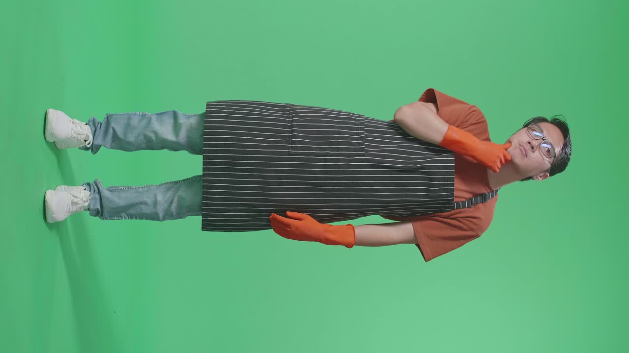 초록색 스크린 스튜디오에 서 있는 동안 무언가에 대해 생각하고 주위를 둘러보는 아프론을 입은 아시아 남성 집주인