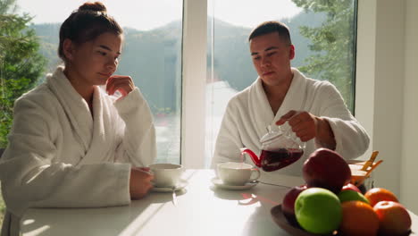 man pours tea into cup sitting with wife at table. young couple spends morning drinking hot tea together in kitchen. concept of warm family relationship