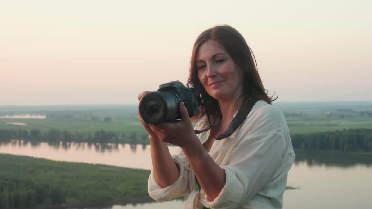 smiling woman in light clothing holding professional camera reviewing shot after capturing sunset over peaceful river landscape with green fields and trees in background