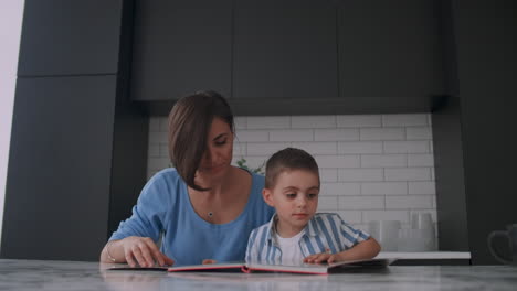 Young-beautiful-mother-and-son-sitting-at-a-table-in-a-bright-kitchen-reading-a-book-and-looking-at-pictures-poking-a-finger-into-a-book-and-leafing-through-the-pages