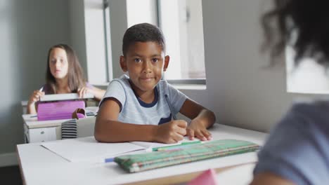 Video-of-happy-biracial-boy-sitting-at-desk-in-classsroom