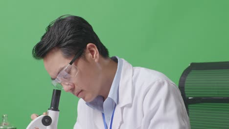 close up of side view of asian man scientist looking through a microscope on the table with test tube then smiling and crossing her arms while sitting in the green screen background laboratory