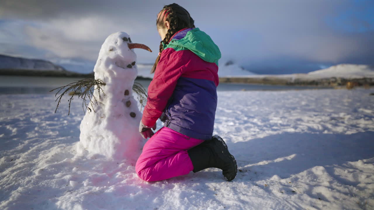 Colorful winter outfit girl building snowman in snowy mountain holiday landscape