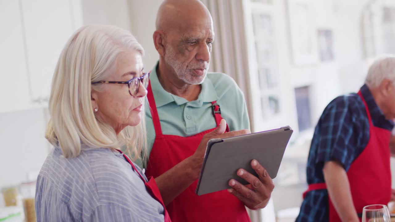 Happy diverse senior male and female friends using tablet, preparing food in kitchen, slow motion