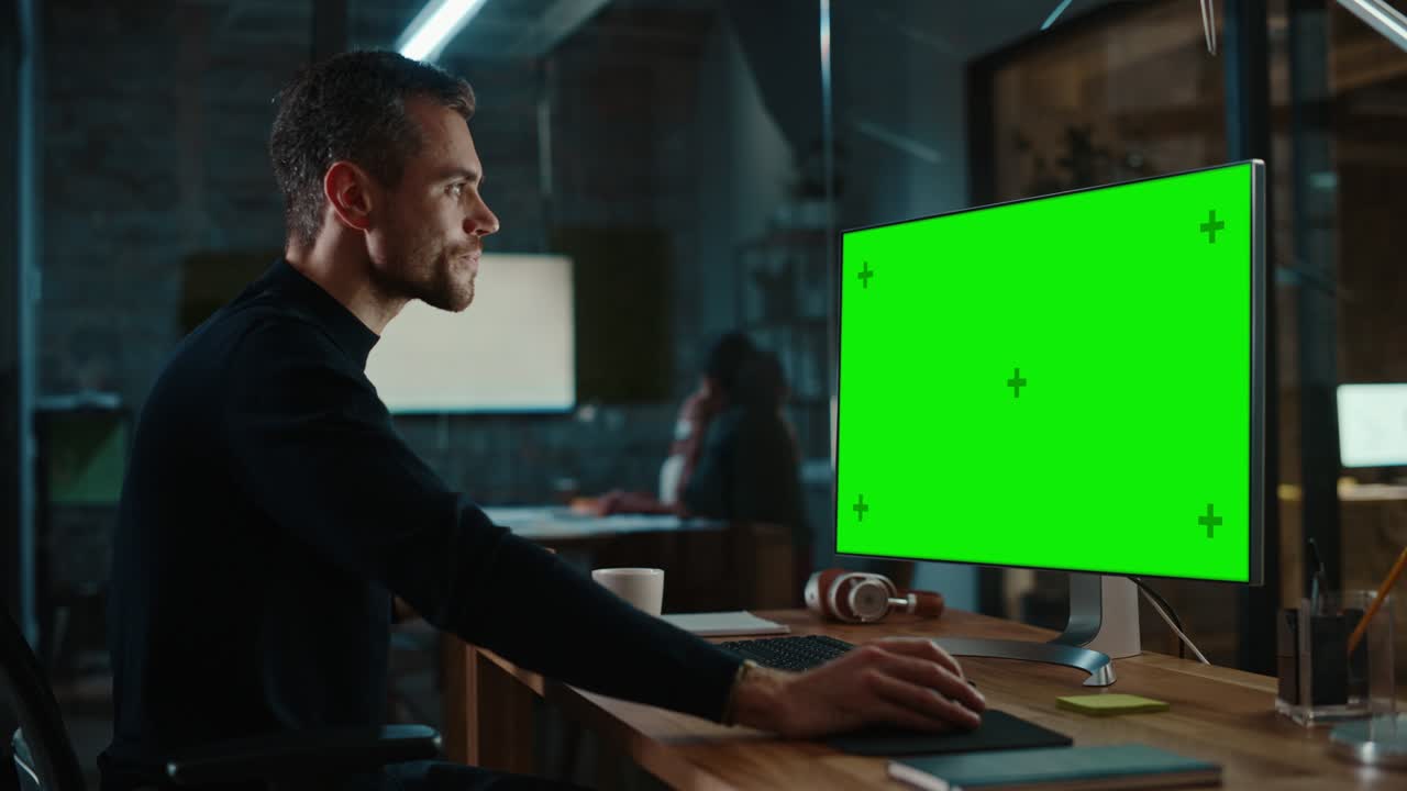 joven especialista guapo trabajando en una computadora de escritorio con pantalla verde simulacro de visualización en la oficina creativa. gerente masculino toma un sorbo de té o café de una taza blanca que recoge de su mesa de trabajo.