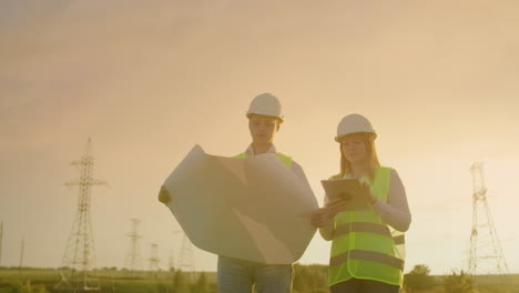 dos electricistas trabajan juntos de pie en el campo cerca de la línea de transmisión de electricidad en cascos. dos electricistas trabajan juntos de pie en el campo cerca con torres de transmisión de energía. combustible ecológico