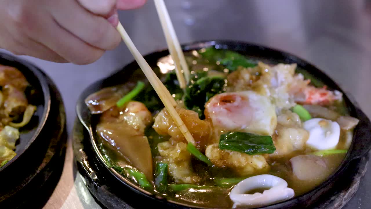 Hand uses chopsticks to pick up seafood pad thai noodles from sizzling hot plate, night market