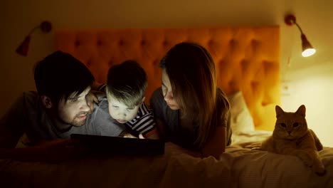 happy family with little son and funny cat lying in bed at home and surfing social media on tablet computer before sleeping