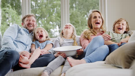family sitting on sofa at home laughing and watching tv with popcorn together