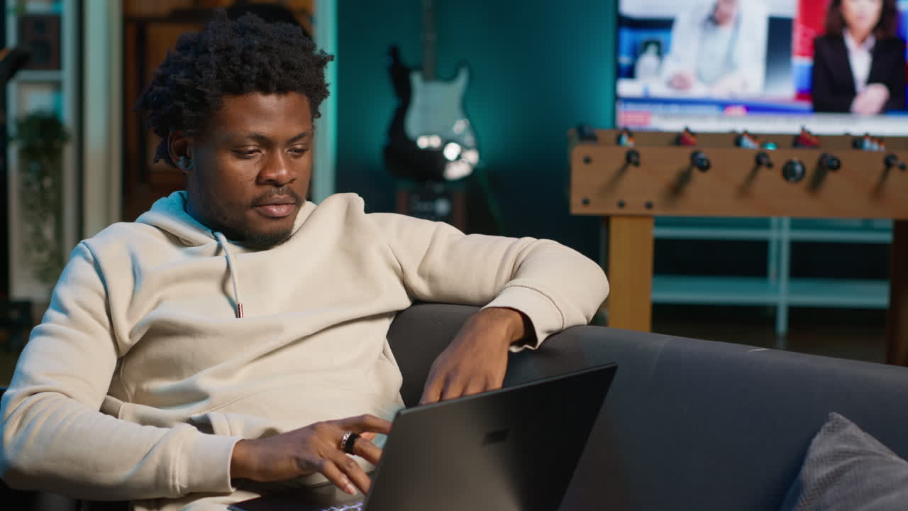 Casual person opening his laptop at home to solve daily tasks on his couch, doing remote freelance work online. Young black man surfing the web multitasking in loft apartment, tech savvy. Camera A.