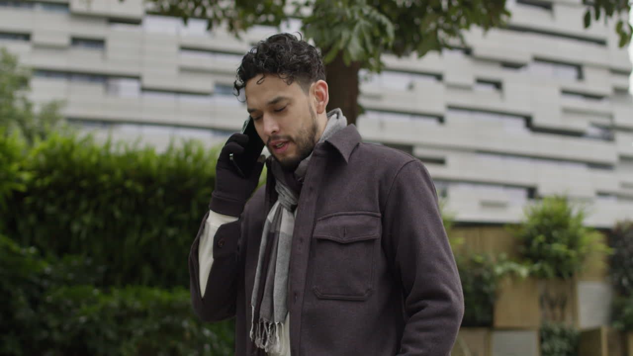 겨울 옷을 입고 장갑과 스카프를 입은 백인 남성이 스마트 시티의 현대적인 주거 지역에서 새로운 스마트폰으로 야외에서 이야기합니다.