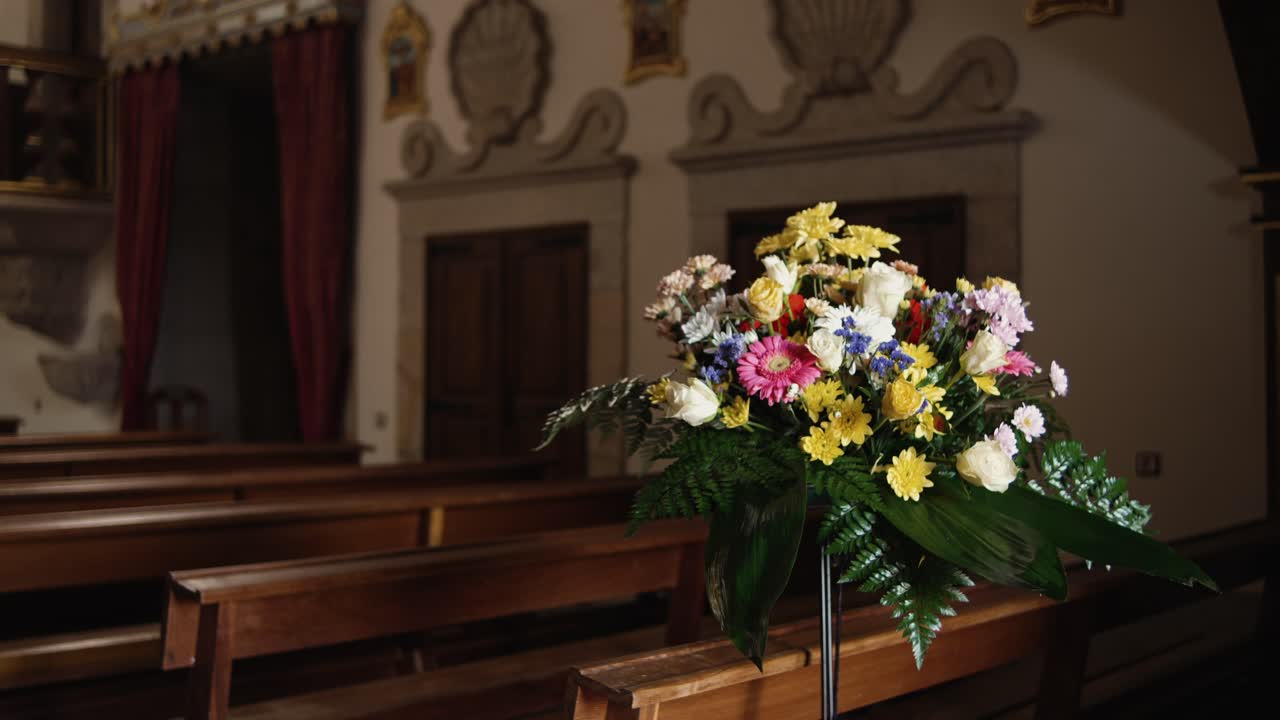 Bouquet di fiori colorati su un supporto all'interno di una chiesa con banchi di legno ed elementi decorativi alle pareti sullo sfondo.