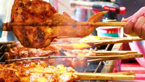 close-up of skewered chicken being grilled, with smoke rising and hands turning the skewers.