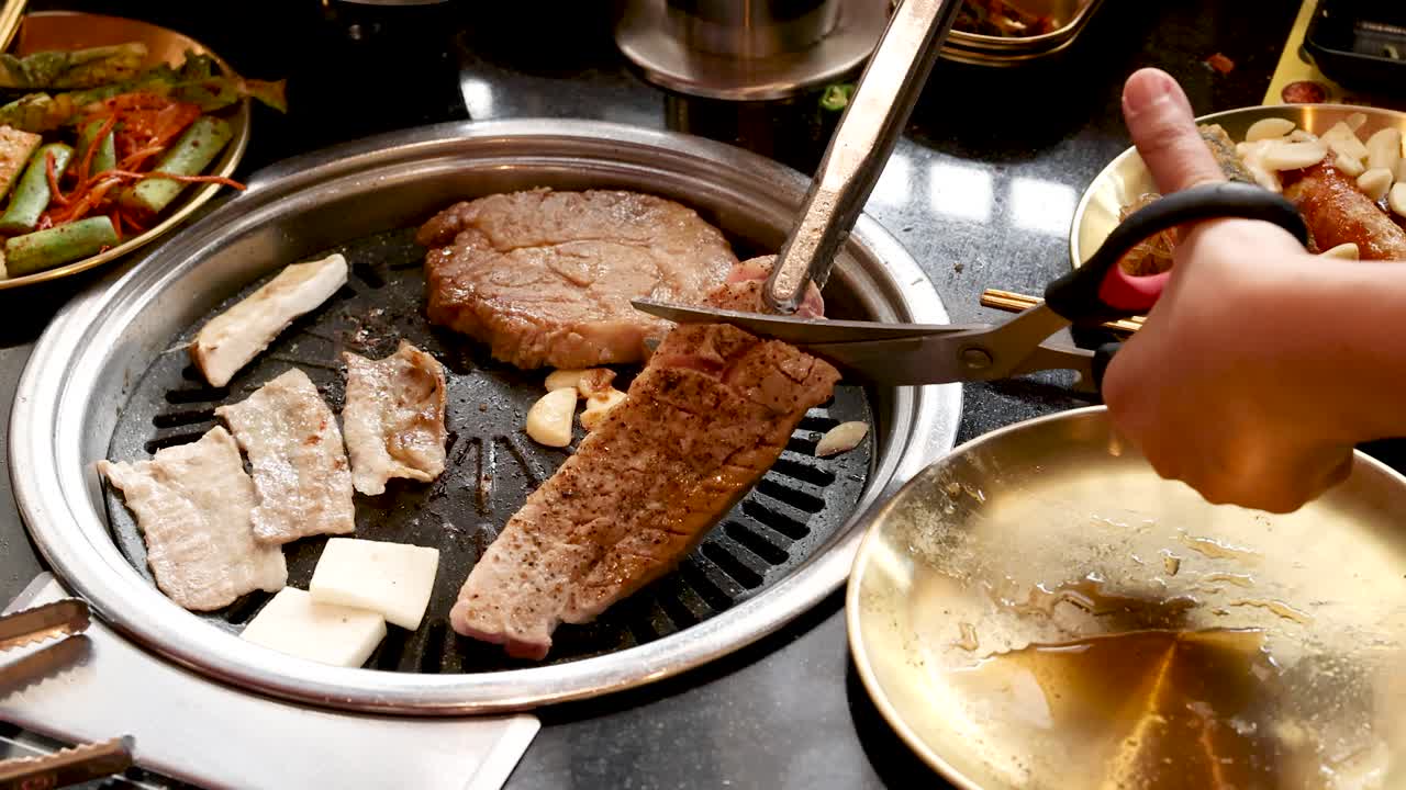 Hand uses scissors to cut pork on tabletop grill, bright lighting, Korean barbecue restaurant setting
