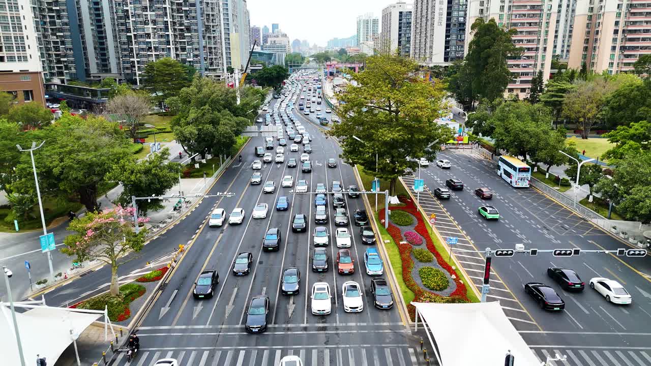 Aerial view of a bustling city street in Shenzhen, China, showcasing heavy traffic, organized lanes, and urban greenery. The scene captures modern infrastructure, daily commute, and urban movement.