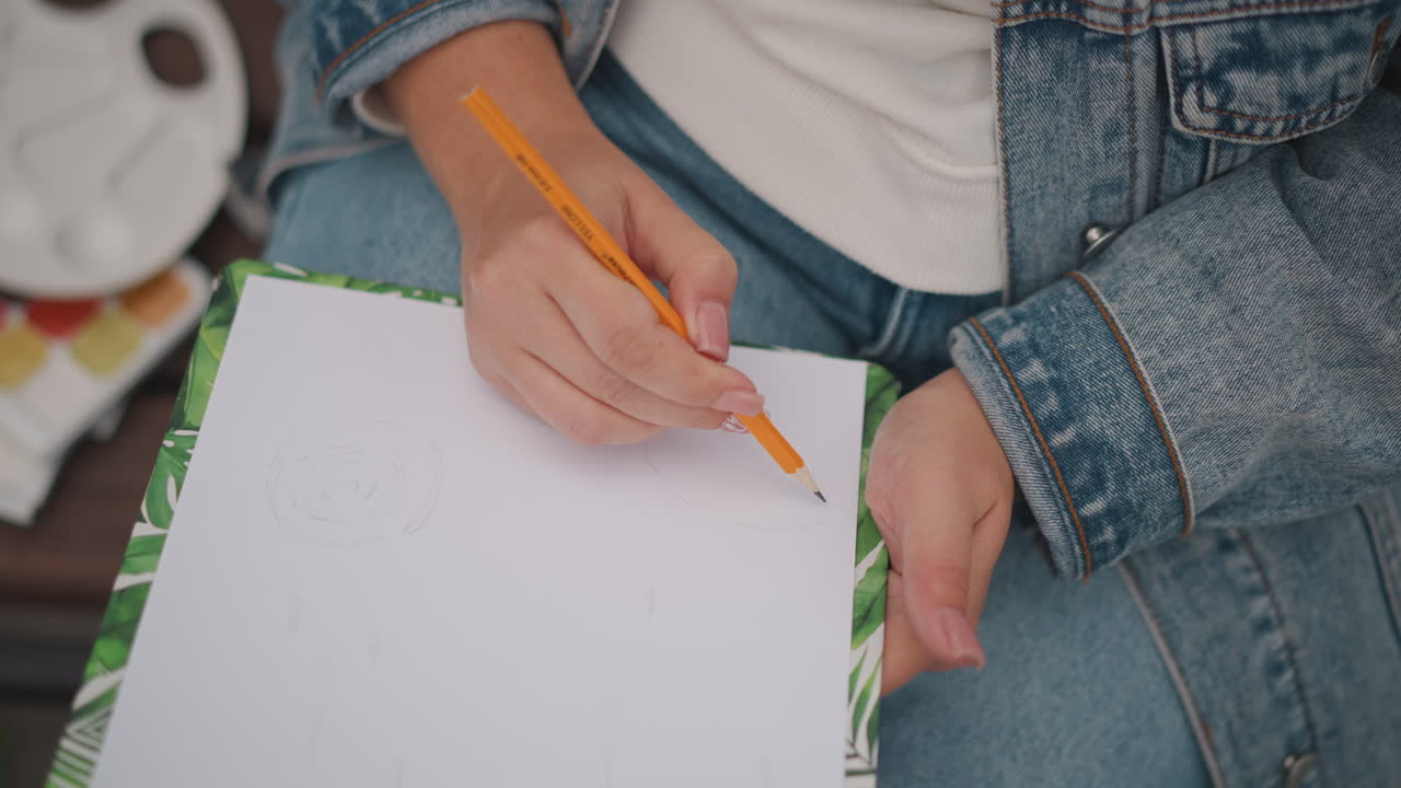 Mani che disegnano su un foglio bianco con una matita, giacca di jeans su una panchina all'aperto, tavolozza di colori visibile, sessione d'arte rilassata, luce naturale e bokeh morbido, pratica amatoriale e ispirazione
