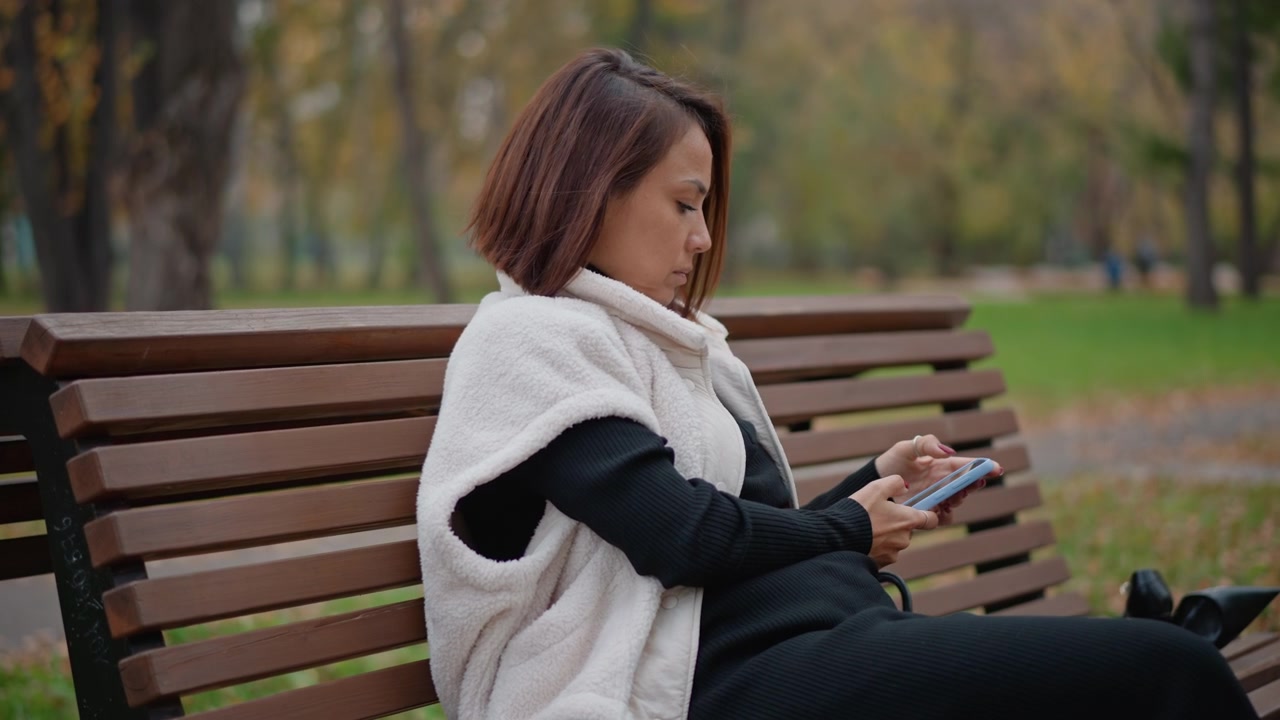 Young woman sits on wooden park bench holding smartphone with both hands, leaning slightly forward with focused expression while enjoying quiet autumn moments beside tree lined path