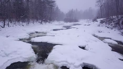 Luftaufnahme-In-Zeitlupe,-Die-Während-Eines-Winterschneesturms-Flussaufwärts-Entlang-Eines-Eisigen-Flusses-Fliegt