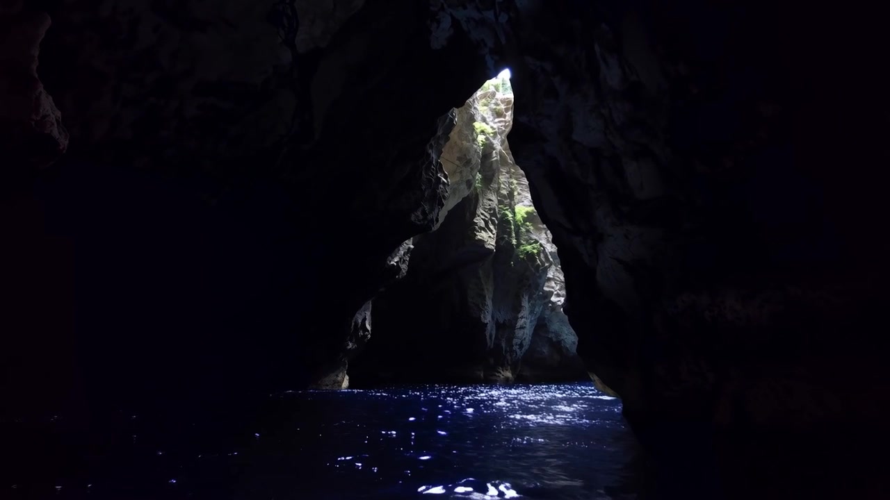 Telecamera in movimento all'interno di una grotta che rivela un ingresso triangolare illuminato, con la luce del sole che cattura il muschio e le increspature dell'acqua.
