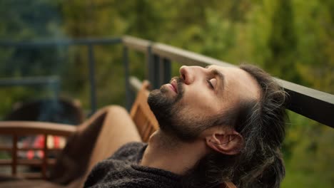 close-up shot: a young brunette man with closed eyes is resting on a sofa on the balcony of a country house overlooking the mountains and coniferous forest. spiritual rest, outdoor leisure