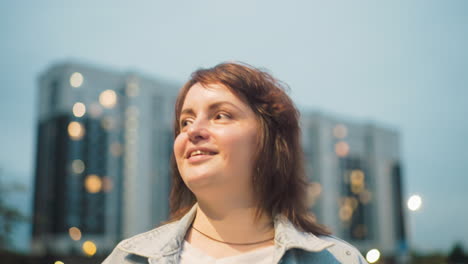 confident woman smiles slightly while standing outdoors in evening near high-rise buildings, wearing denim jacket under soft sky, radiating warmth, independence, and peace in urban lifestyle