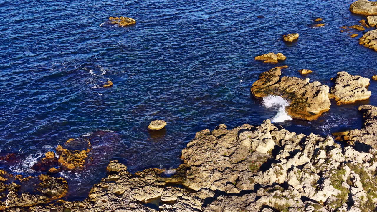 Serene coastal landscape with rocky formations and gentle waves under a bright, clear sky at midday near the seashore