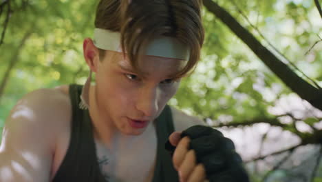 close up view white man wearing headband intense focused eyes bouncing gloved palms off face guard amid blurred green foliage background conveying disciplined martial training moment