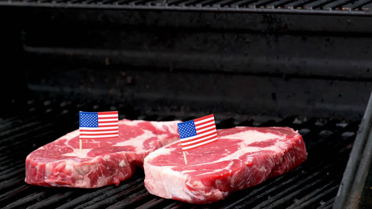 그릴에 앉아 손으로 요리하는 두 개의 육즙이 많은 립아이 스테이크가 들어와 이쑤시개로 작은 미국 국기를 그 중 하나에 꽂습니다.
