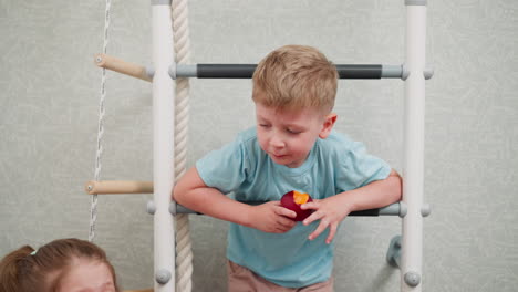 little boy hanging over indoor ladder eating juicy mango while little girl sits nearby, vibrant playful atmosphere, casual home setting with soft pastel background