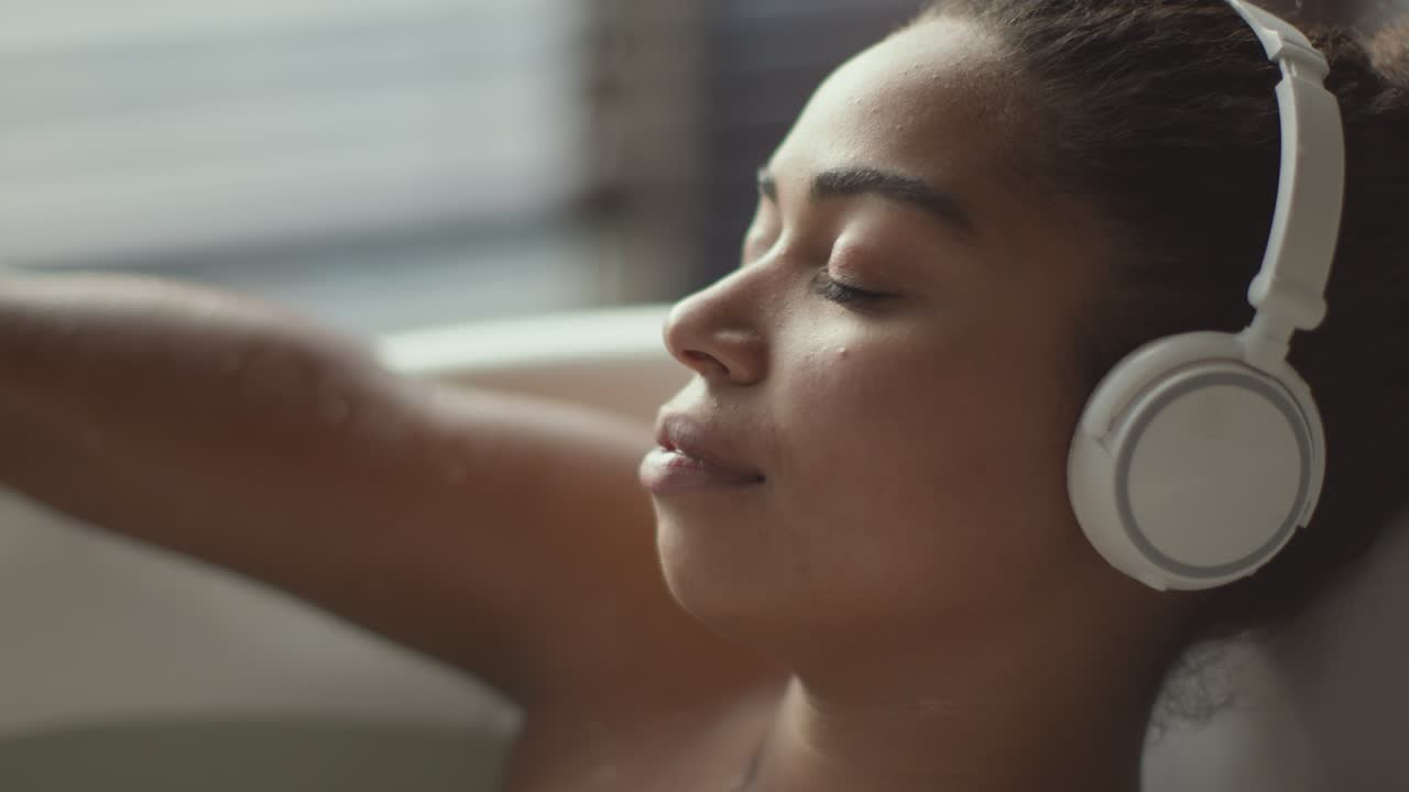 젊은 아프리카계 미국인 여성이 무선 헤드을 통해 음악을 듣고 눈을 감고 뜨거운 목욕탕에서 휴식을 취하고 있습니다.