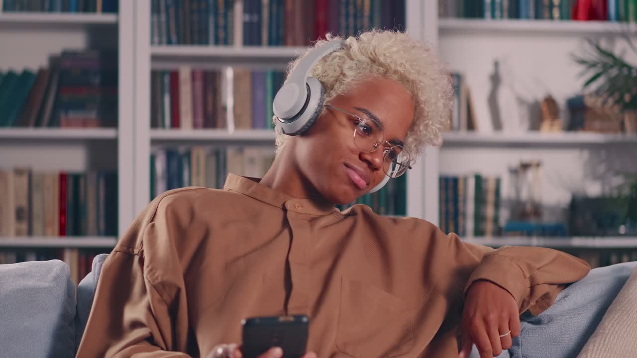 African woman relaxing on sofa holds smartphone listens song through headphones