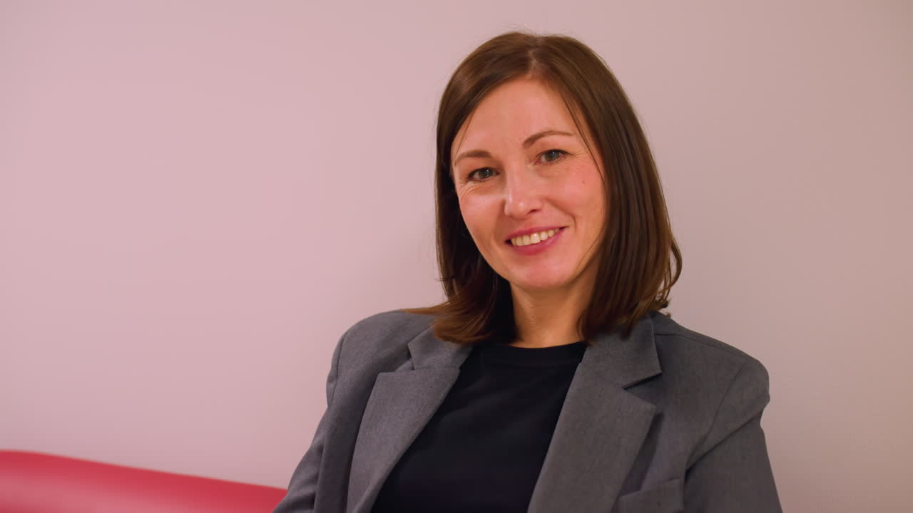 Imprenditrice sorridente e sicura di sé seduta su un divano rosso nel corridoio di un ufficio moderno. Atmosfera rilassata ma professionale, che guarda direttamente la fotocamera, a rappresentare un ambiente di lavoro positivo.