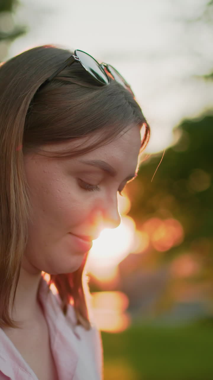 joven con camiseta rosa con gafas en la cabeza, con una cálida sonrisa, mira hacia abajo pensativa, bañada en la luz del sol, con llamaradas solares y vegetación borrosa en el fondo